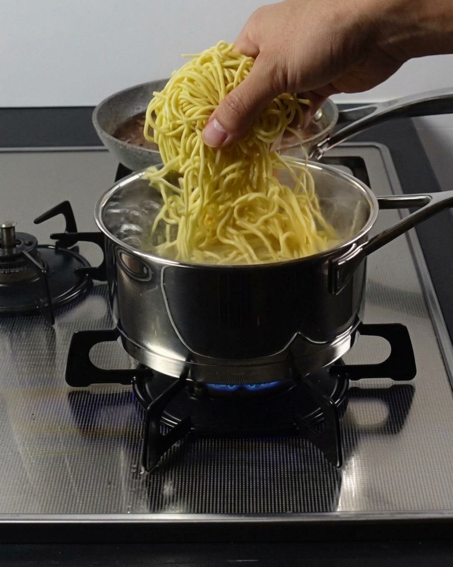 A hand lowering a bundle of thick, yellow ramen noodles into a stainless steel pot of boiling water on a gas stove.