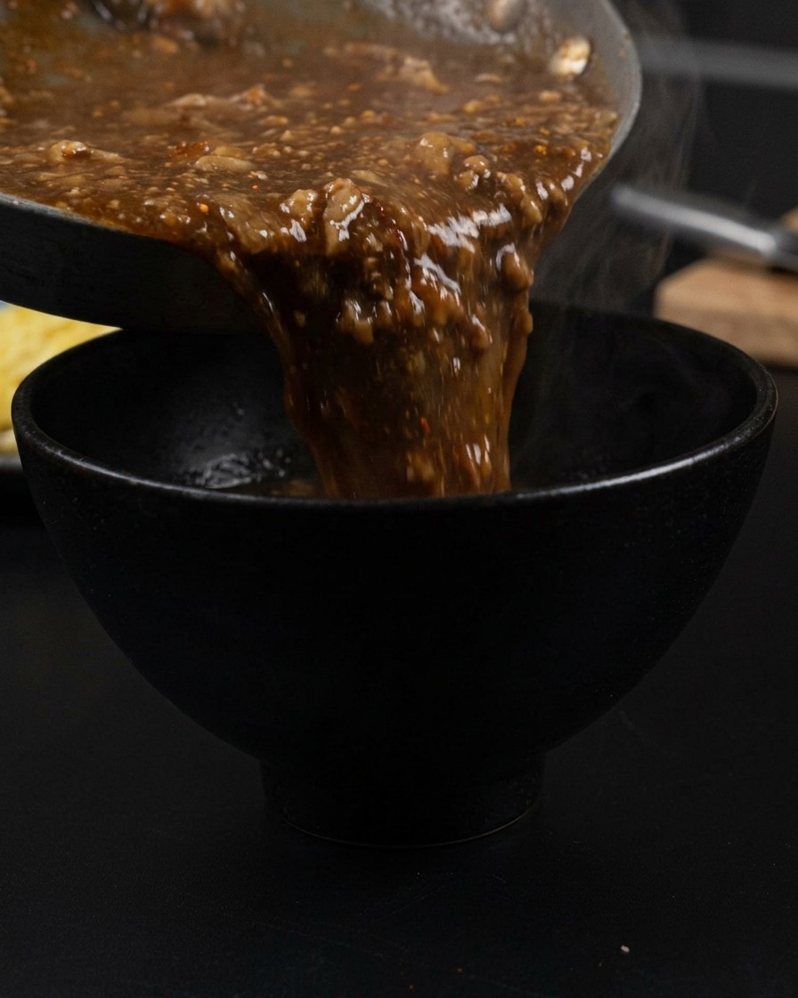 Thick, dark brown savory broth with visible pieces of pork and aromatics being poured from a pan into a matte black bowl, with steam rising from the liquid.
