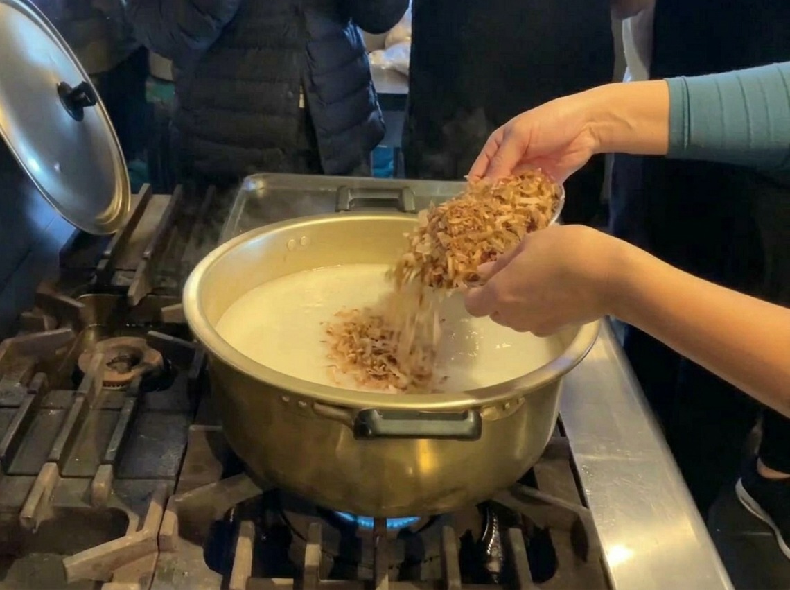 A person pouring a large quantity of brown dried fish flakes from a silver bowl into a large golden pot of white broth.