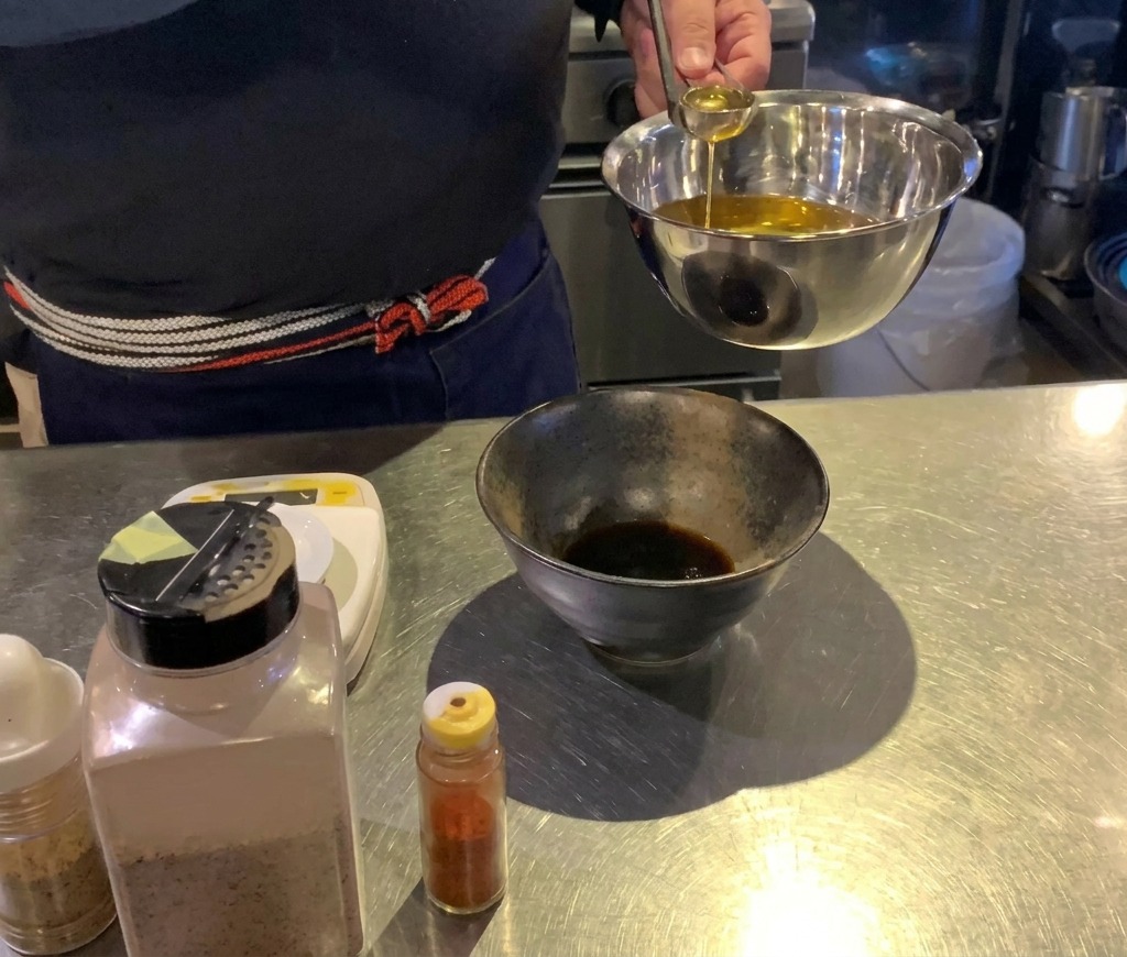 A cook using a small silver ladle to pour golden oil into a black bowl containing a dark sauce.