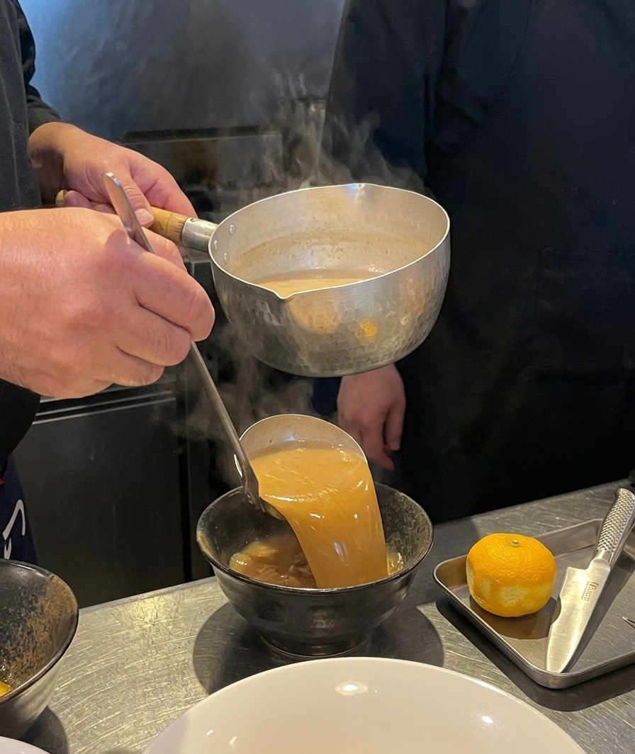 Thick brown broth being poured from a ladle into a black ceramic bowl.
