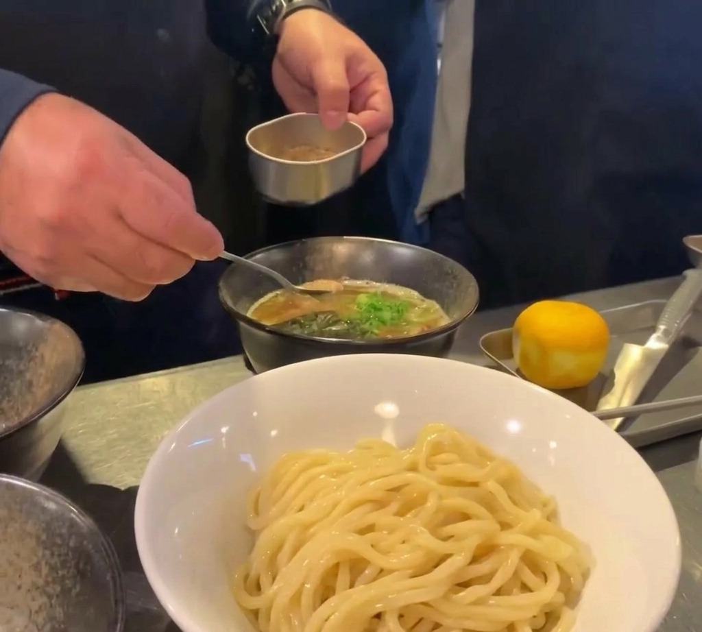 A small spoon depositing a mound of brown fish powder into a bowl of dark broth garnished with green onions.