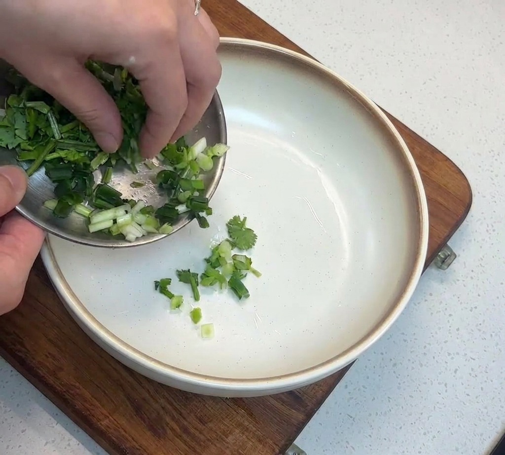 A close-up shot of chopped green scallions being transferred from a small metal dish into a deep white bowl sitting on a wooden cutting board.