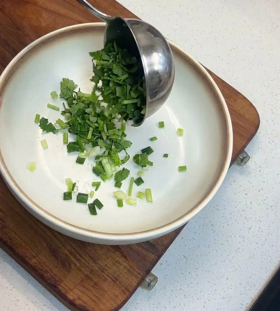 A metal ladle pouring a generous amount of chopped green cilantro and scallions into a shallow white bowl with a brown rim.