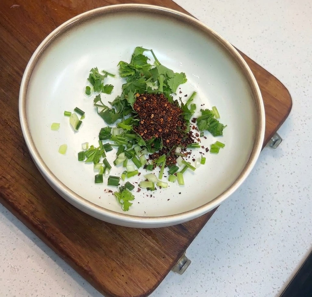A mound of dark red roasted chili flakes sitting atop chopped green herbs in a white ceramic bowl.