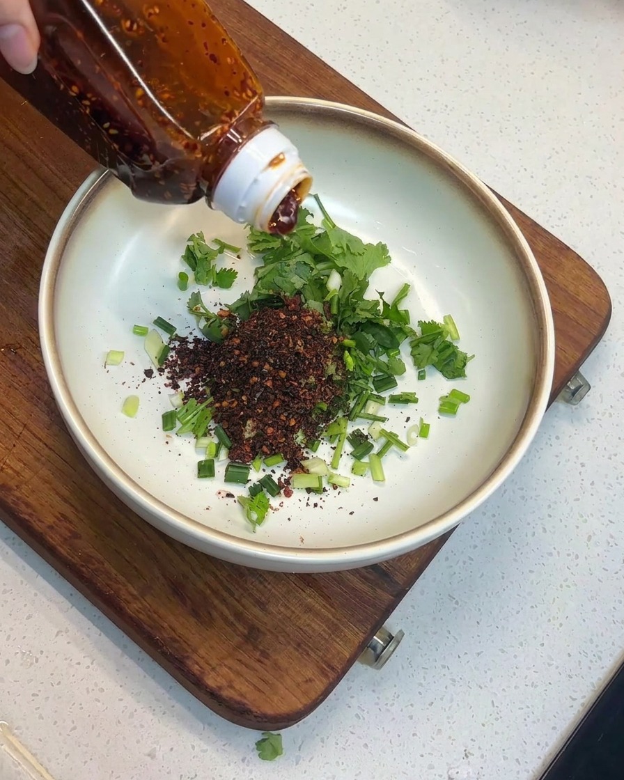 Red chili oil being poured from a clear plastic bottle onto a pile of chili flakes and herbs in a bowl.
