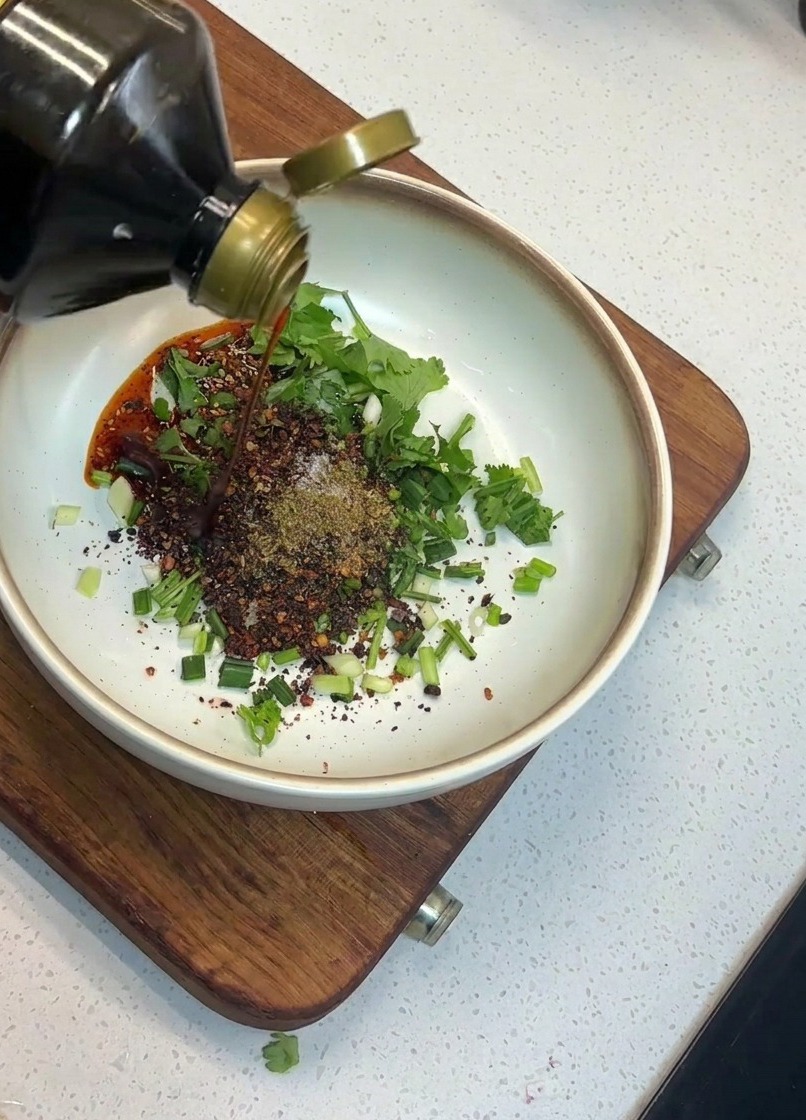 A hand pouring dark soy sauce from a glass bottle into a white bowl containing chopped scallions, cilantro, and red chili flakes.
