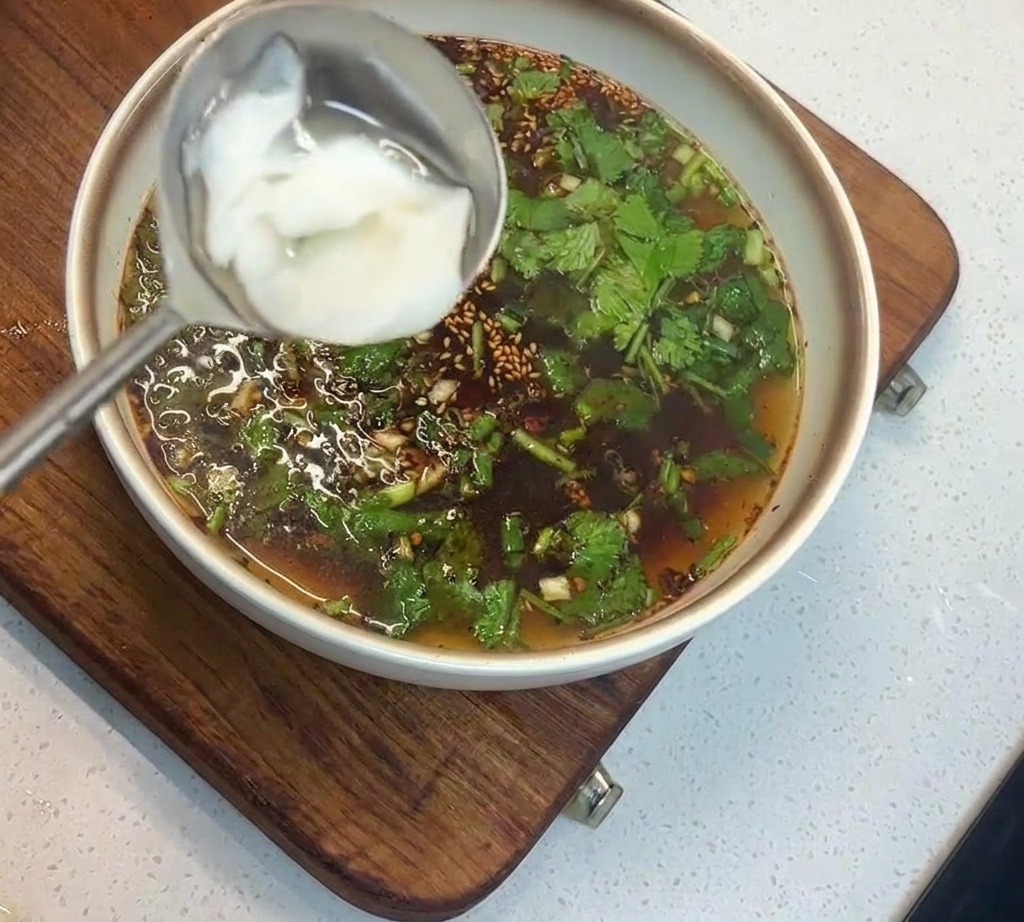 A metal ladle holding a scoop of white lard being lowered into a bowl of dark, herb-filled broth.