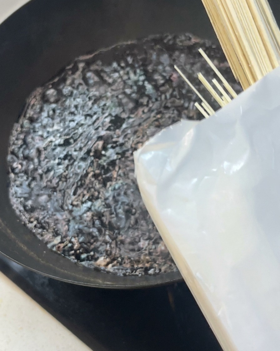 A bundle of straight, dry white noodles being placed into a black wok with bubbling boiling water.