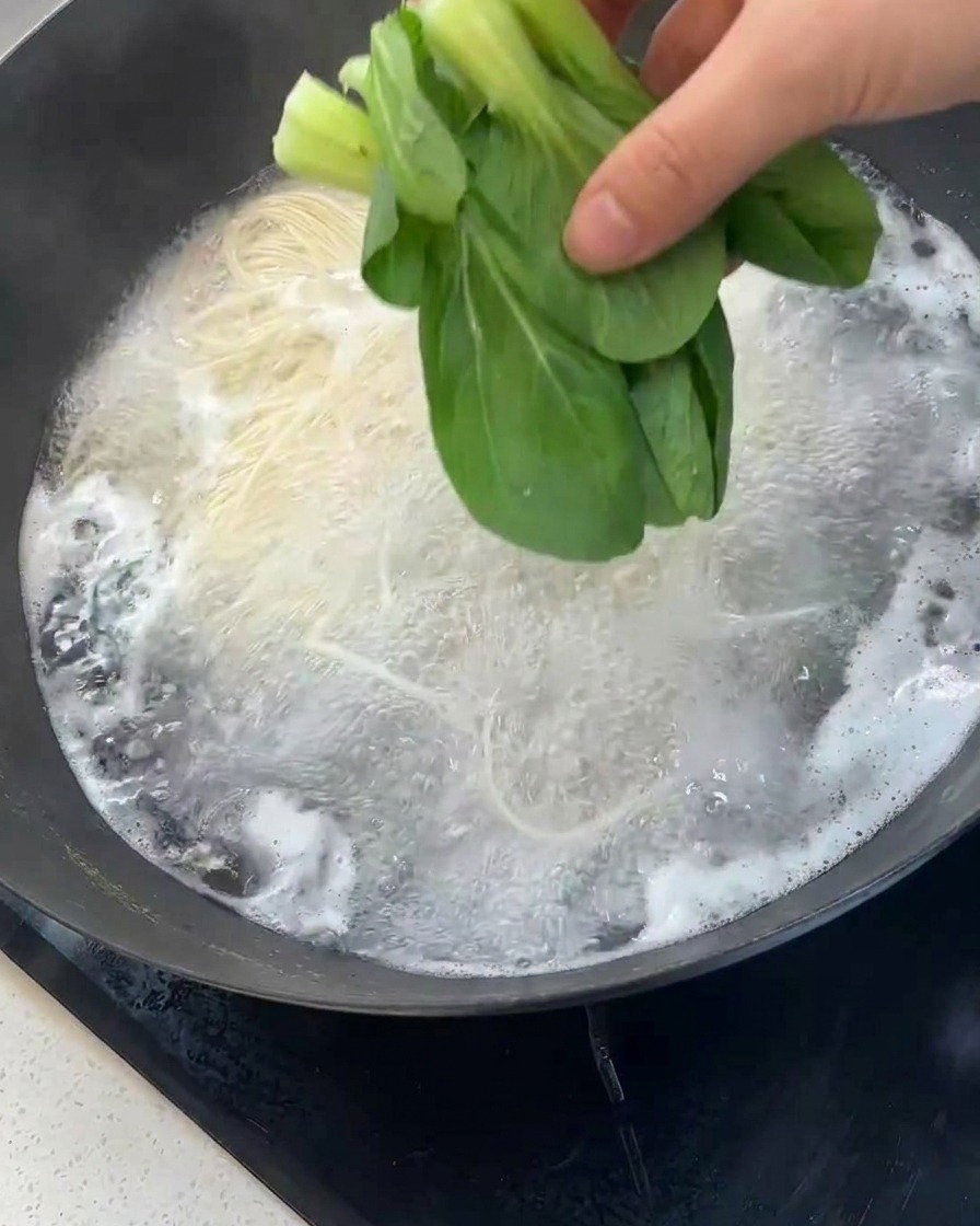 A hand dropping fresh green bok choy into a wok of boiling water filled with white noodles.