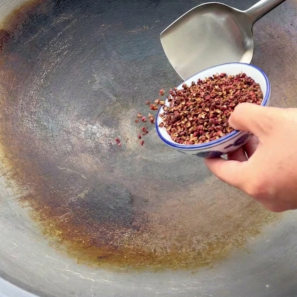 Sichuan peppercorns being poured from a small blue and white bowl into a large seasoned wok for toasting.