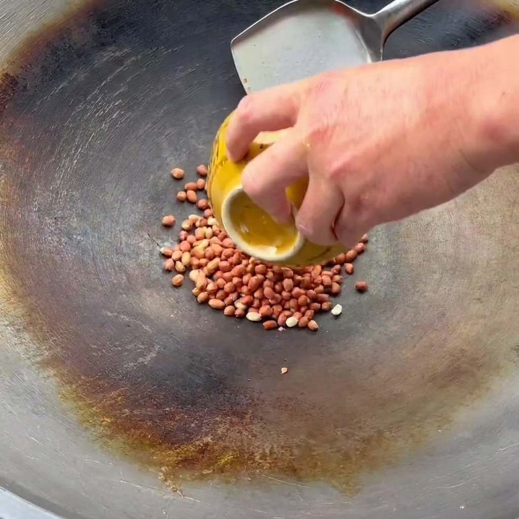 A hand pouring raw peanuts from a yellow bowl into a wok where a metal spatula is ready to stir-fry them.