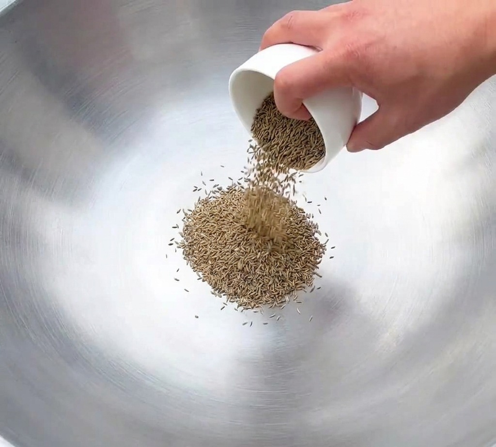 A metal spatula stir-frying a pile of light brown cumin and fennel seeds in the center of a dark, seasoned wok.