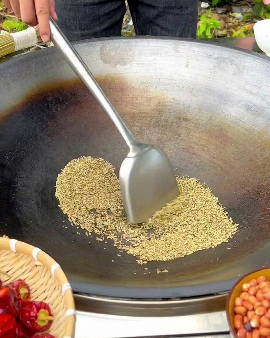 Fennel seeds being stirred with a metal spatula in a large, seasoned iron wok over a low flame.