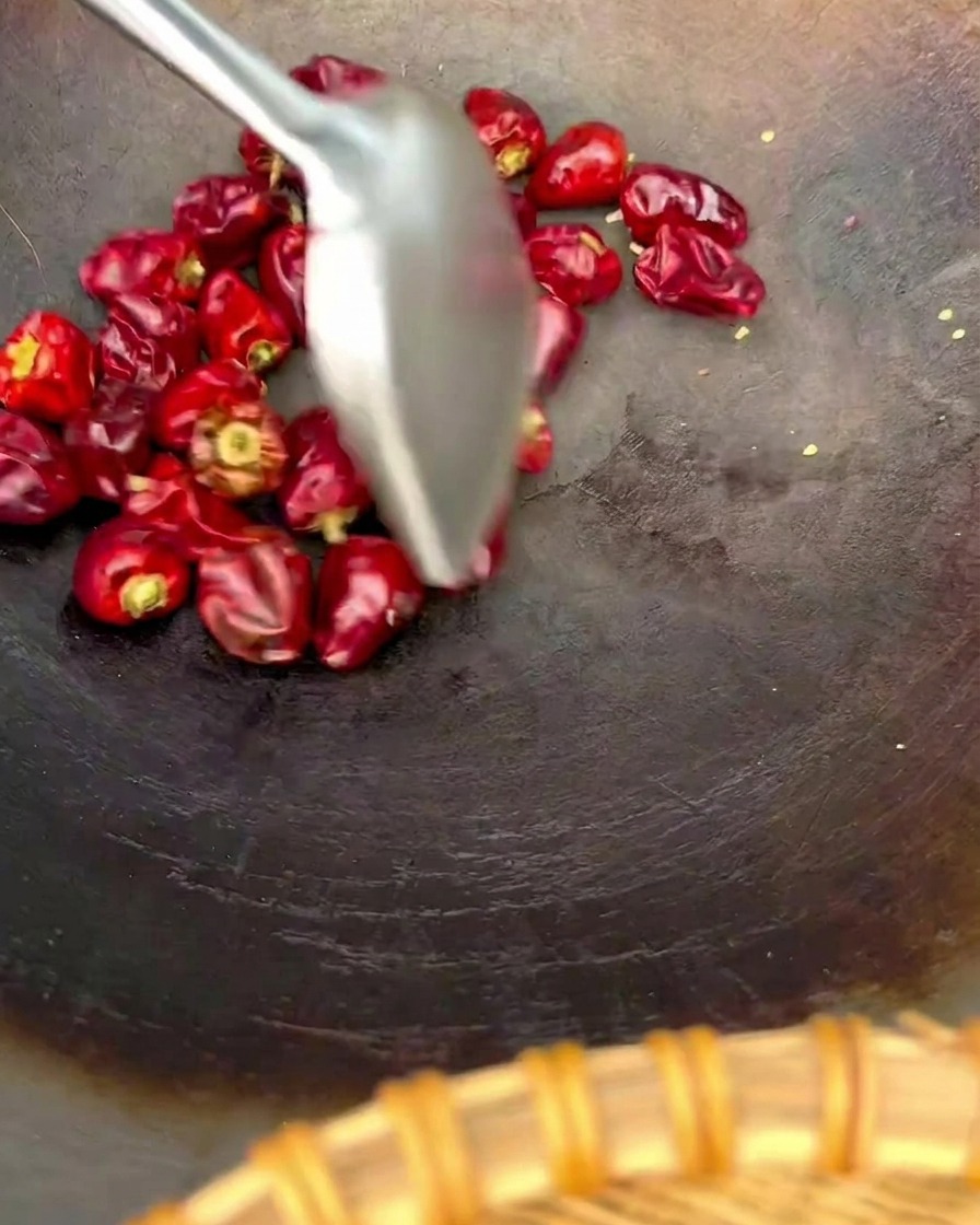 A metal spatula stirring a handful of round red lantern chilies in a dark wok.