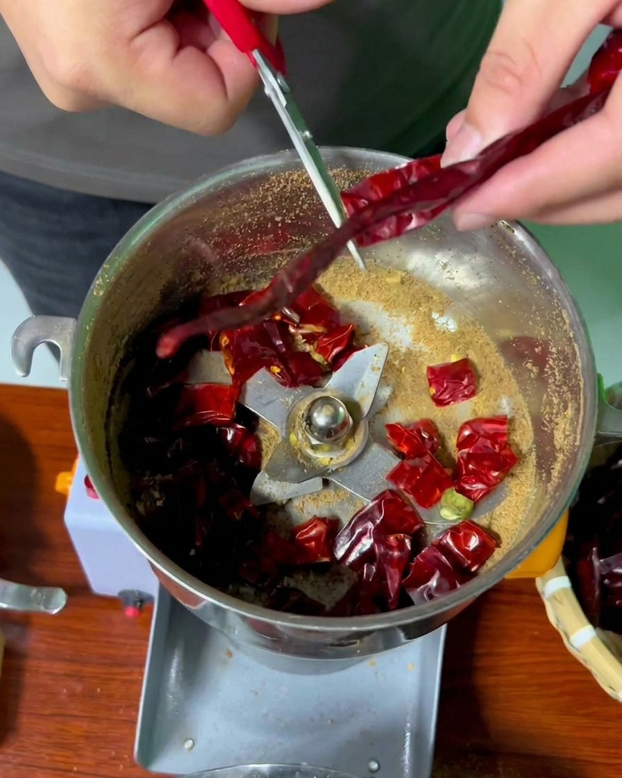Hands using red-handled scissors to snip long dried red chilies into a stainless steel grinder bowl.