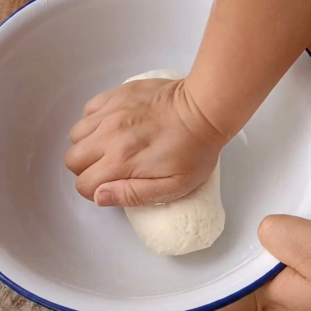 Two hands firmly kneading a ball of dough inside a white mixing bowl.