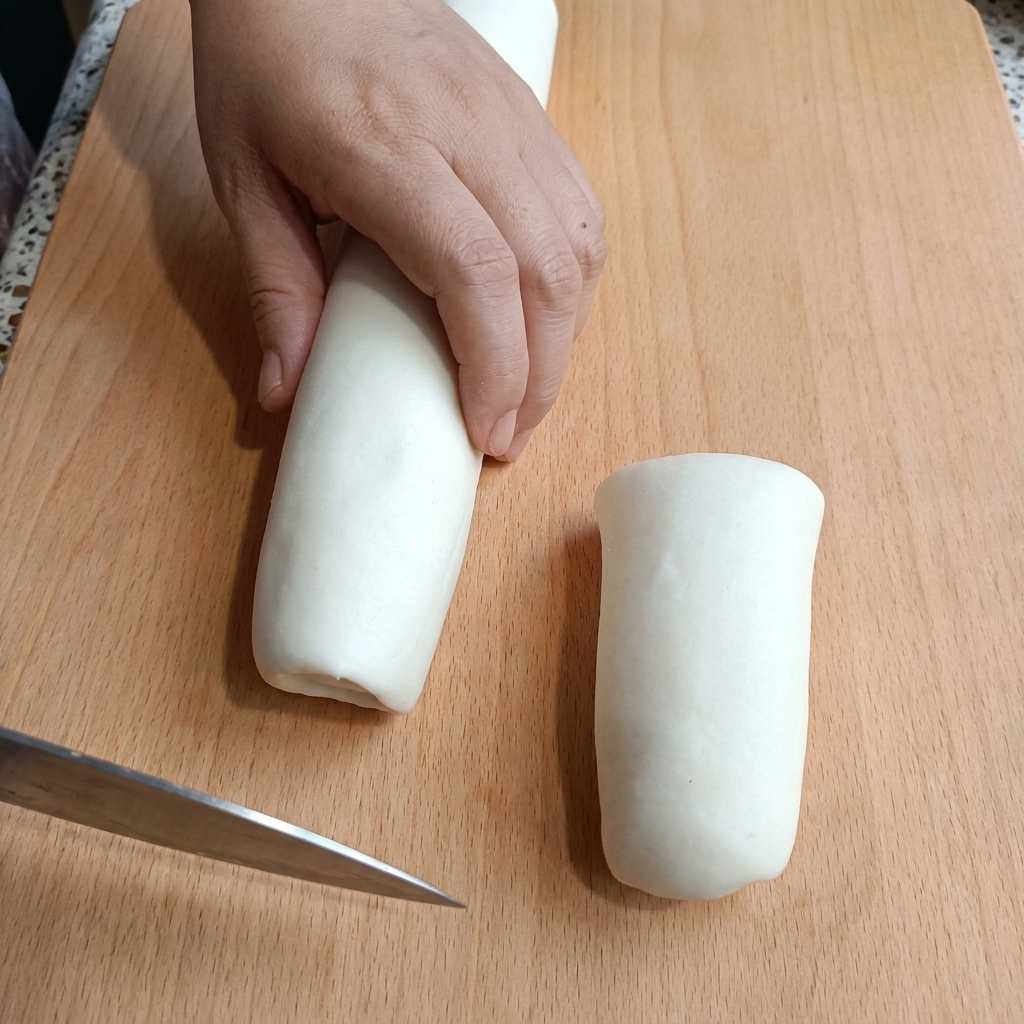 A hand holding a cylinder of white dough on a wooden cutting board while slicing it with a knife.