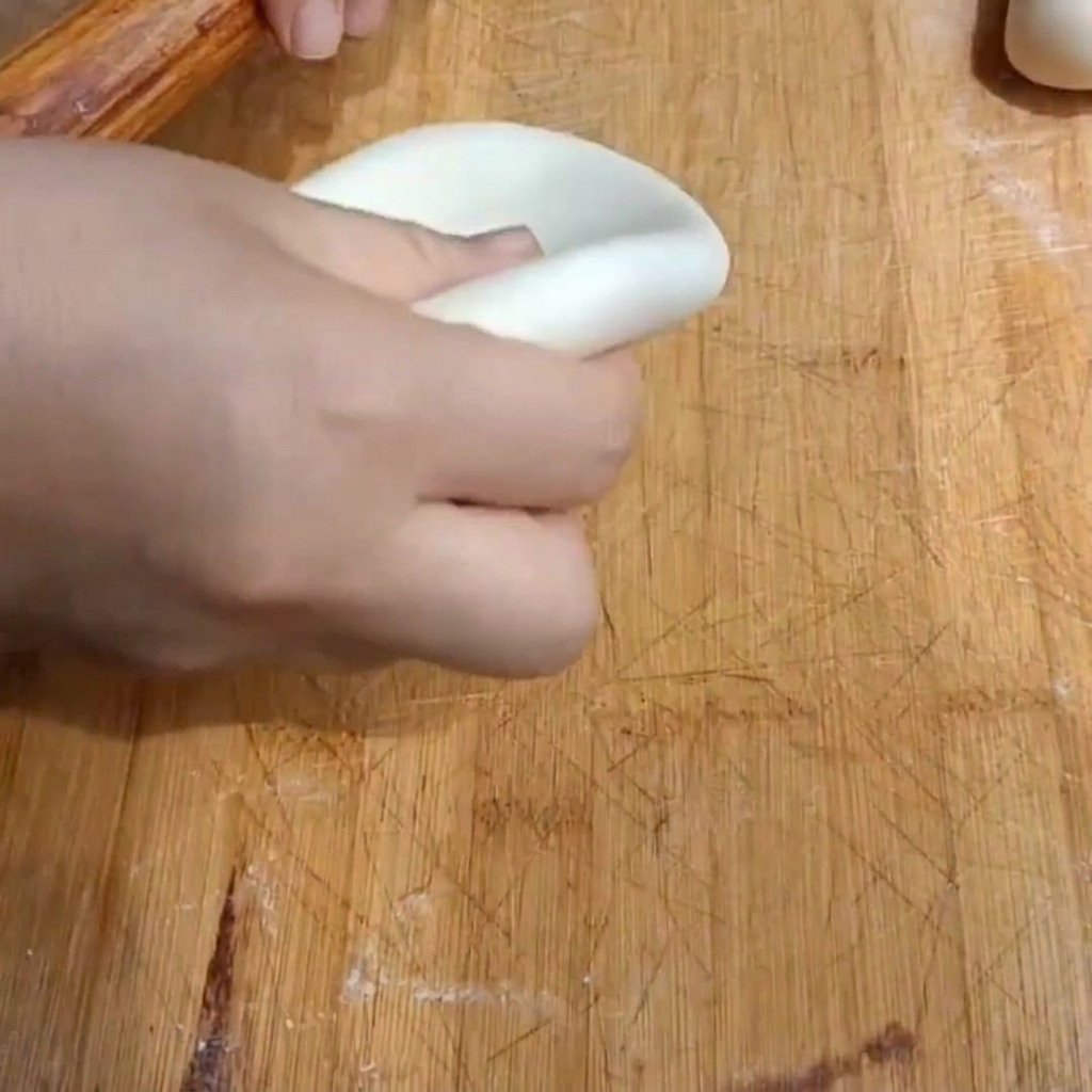 A hand using a wooden rolling pin to flatten a piece of dough into a thick disk on a wooden board.