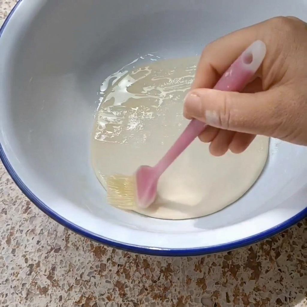 A hand using a silicone brush to coat a round dough disk with vegetable oil inside a blue-rimmed white bowl.