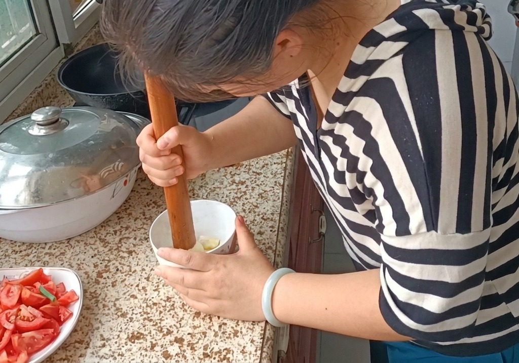 A person using a wooden pestle to pound garlic cloves into a smooth paste in a small white bowl on a granite countertop.