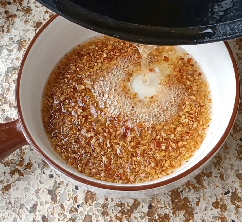 Hot oil being poured from a black pan into a white bowl containing garlic and chili flakes, creating bubbles and foam.