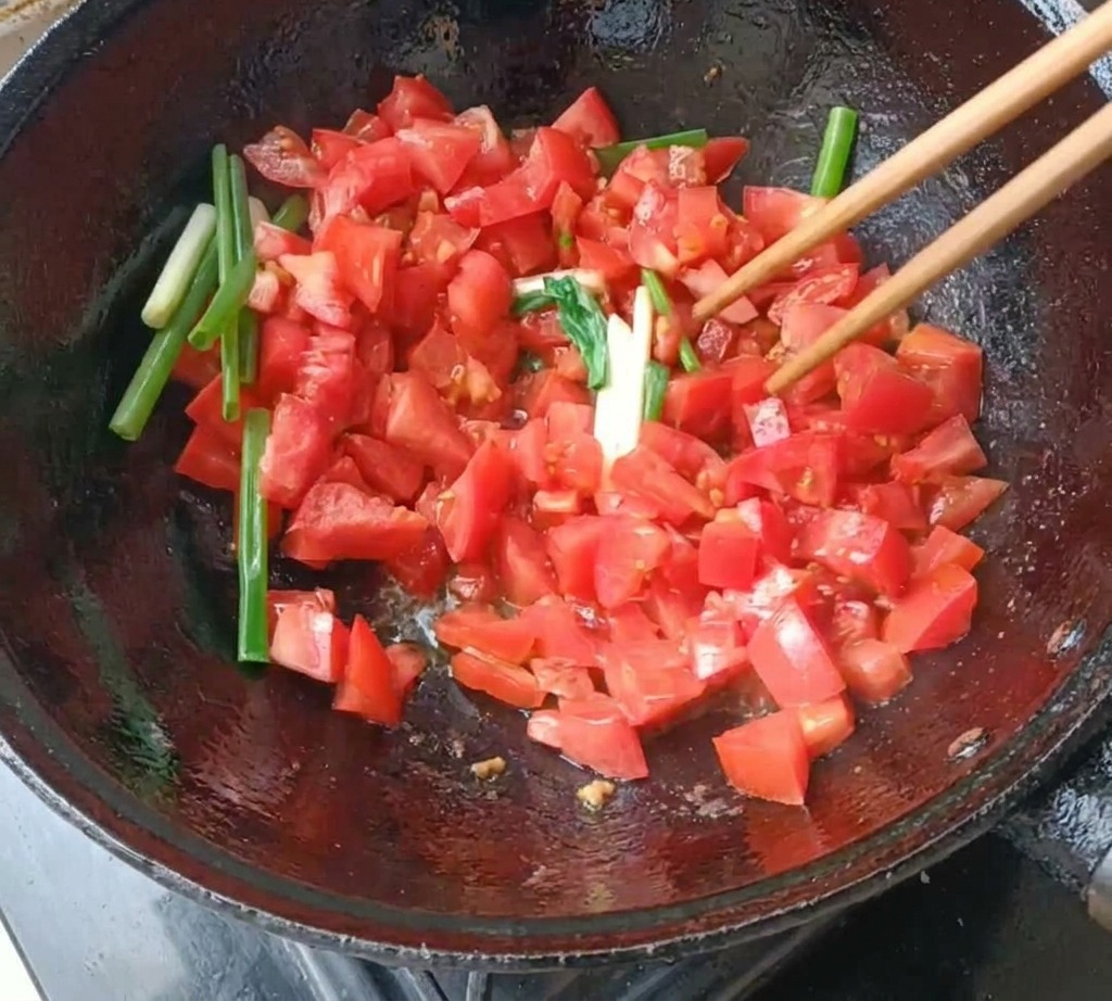 Diced tomatoes and long green onion segments being stir-fried with wooden chopsticks in a dark cast-iron wok.