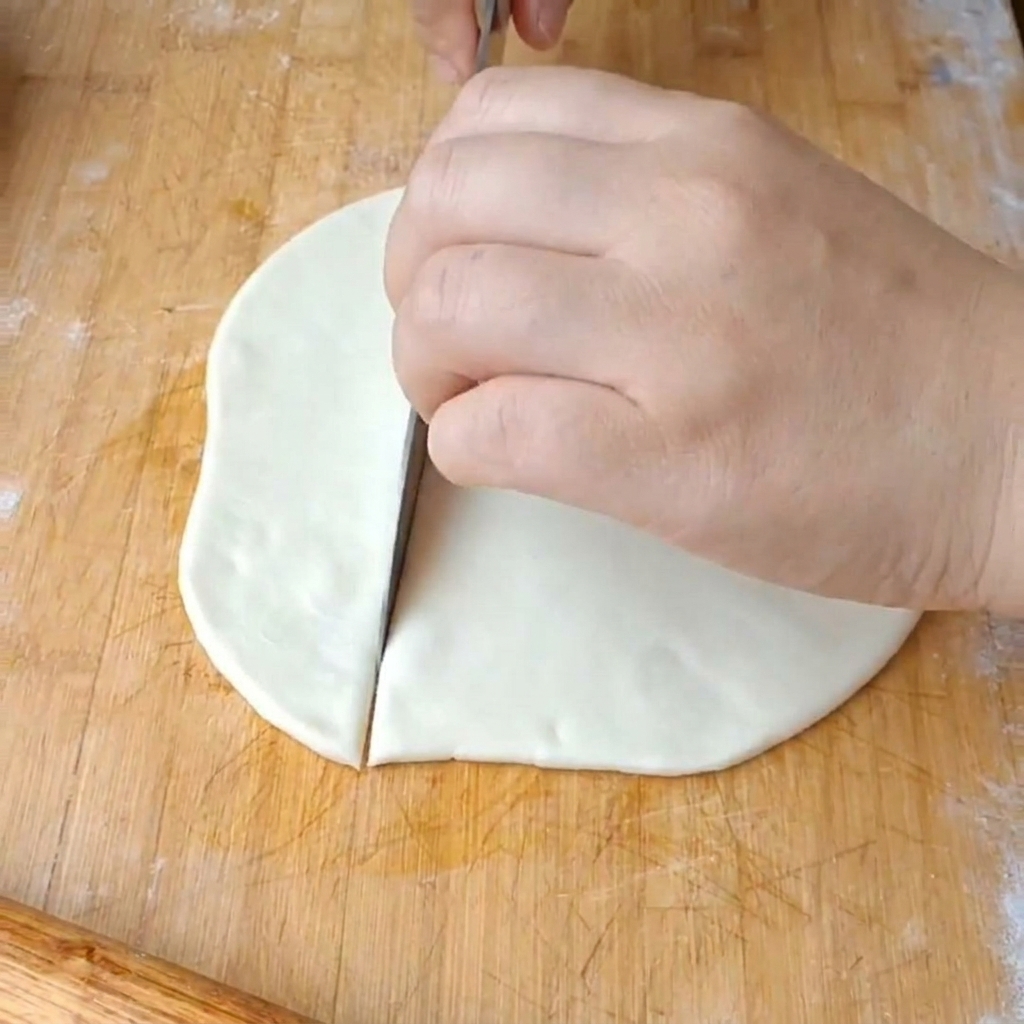 A hand using a large metal knife to cut a flat, white dough disk into wide ribbons on a wooden cutting board.