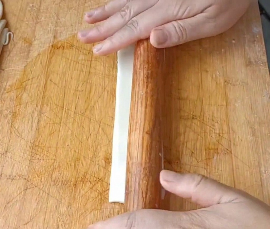 A close-up view of hands using a wooden rolling pin to flatten a long, white strip of oiled dough on a wooden cutting board.