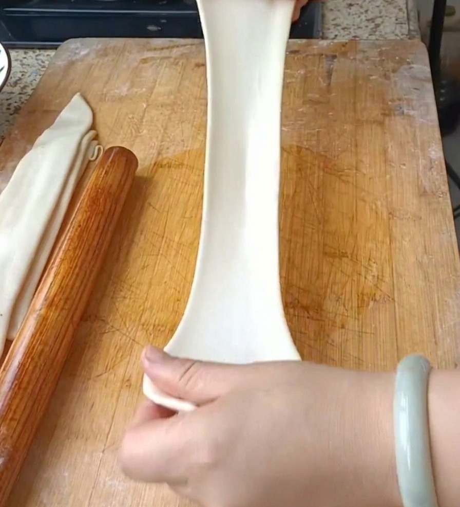 Two hands pulling a wide, translucent white dough strip vertically to form a long noodle over a wooden cutting board.