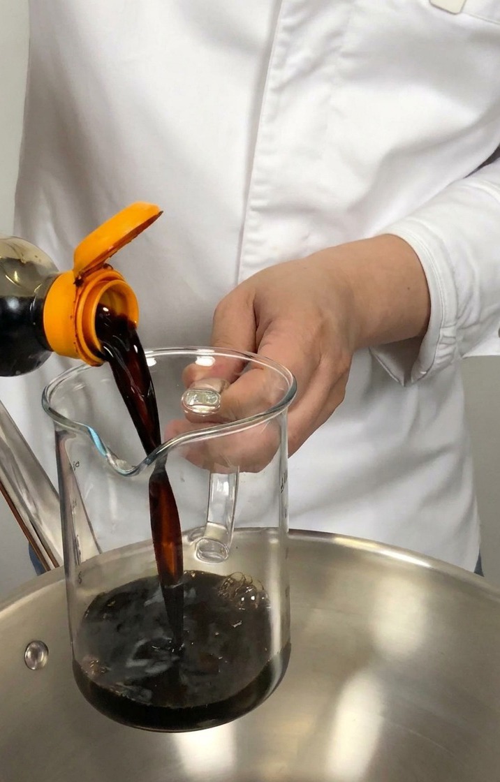 Dark soy sauce being poured from a bottle into a glass measuring cup held over a stainless steel pan.