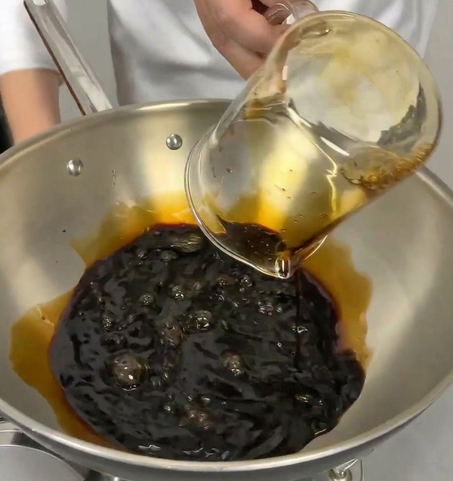 A glass measuring cup pouring clear Japanese cooking wine into a pan containing dark soy sauce.