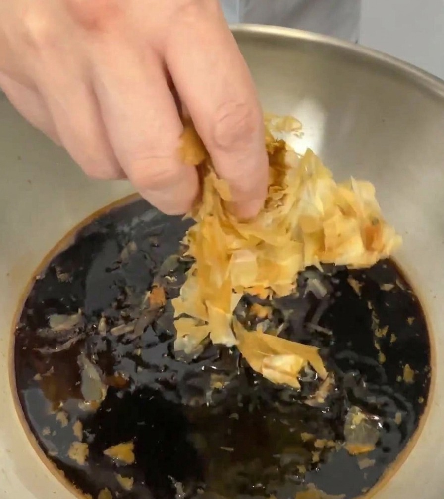 A hand dropping light-colored, thin dried bonito flakes into the dark liquid in the pan.