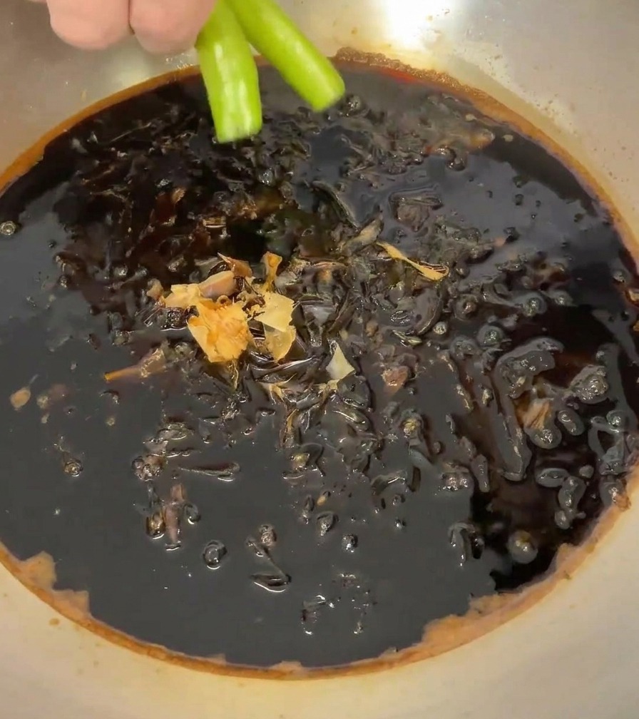 A hand placing two bright green chili peppers into the pan filled with the dark sauce and bonito flakes.