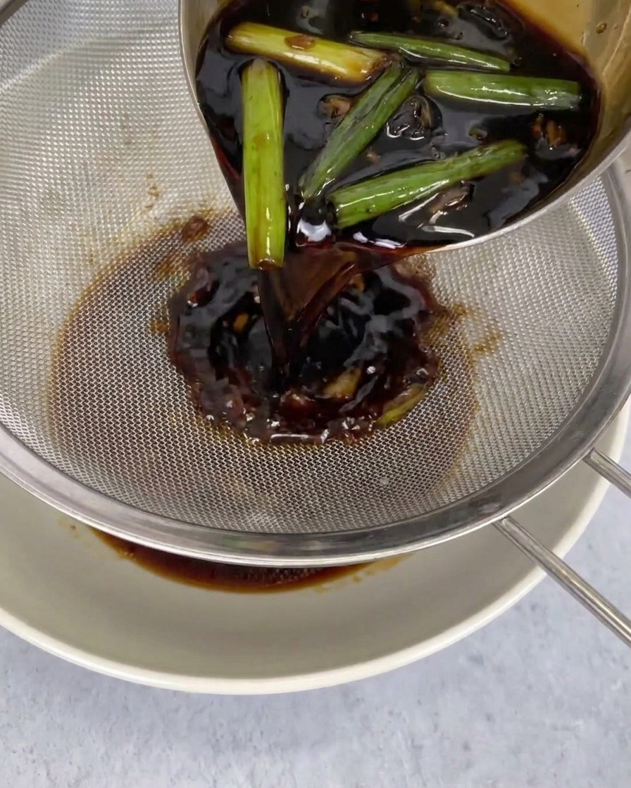 A dark dipping sauce being poured from a ladle through a metal sieve into a white bowl, with green chili peppers and other aromatics being caught in the mesh.