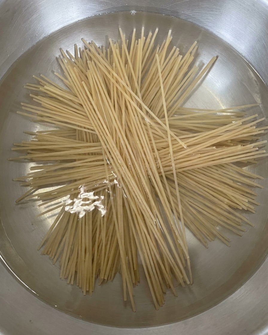 Dry, straight buckwheat noodles being added to a stainless steel pot of boiling water, spreading out in a star pattern.