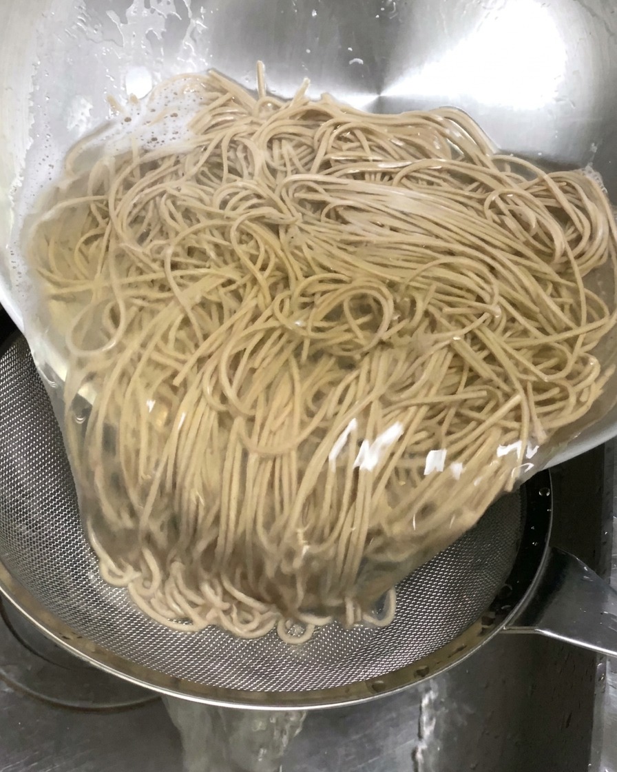 Cooked buckwheat noodles being poured from a large bowl into a metal sieve placed in a sink to drain the liquid.