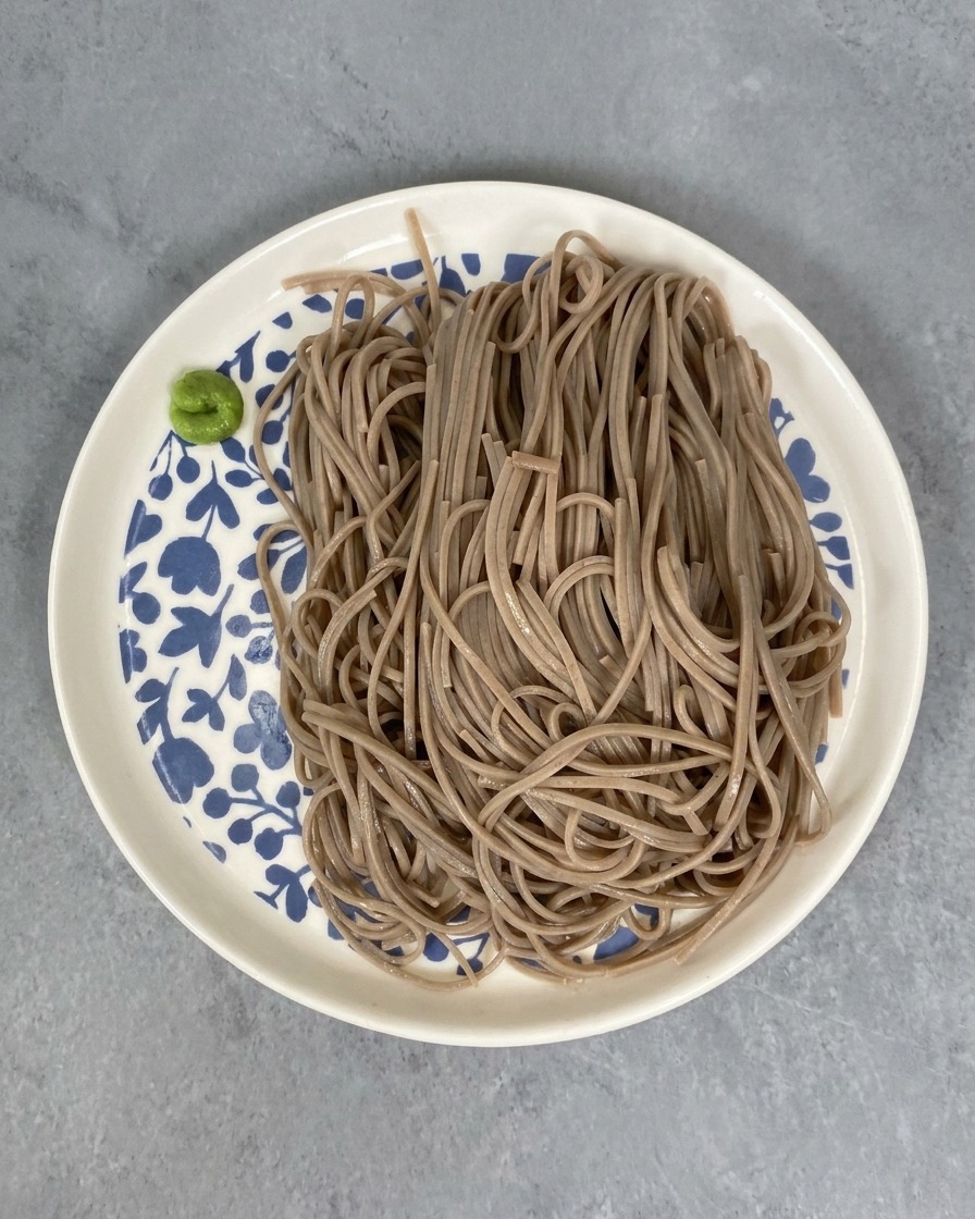 A serving of cooked buckwheat noodles on a white plate with blue floral patterns, accompanied by a small portion of bright green wasabi paste on the side.