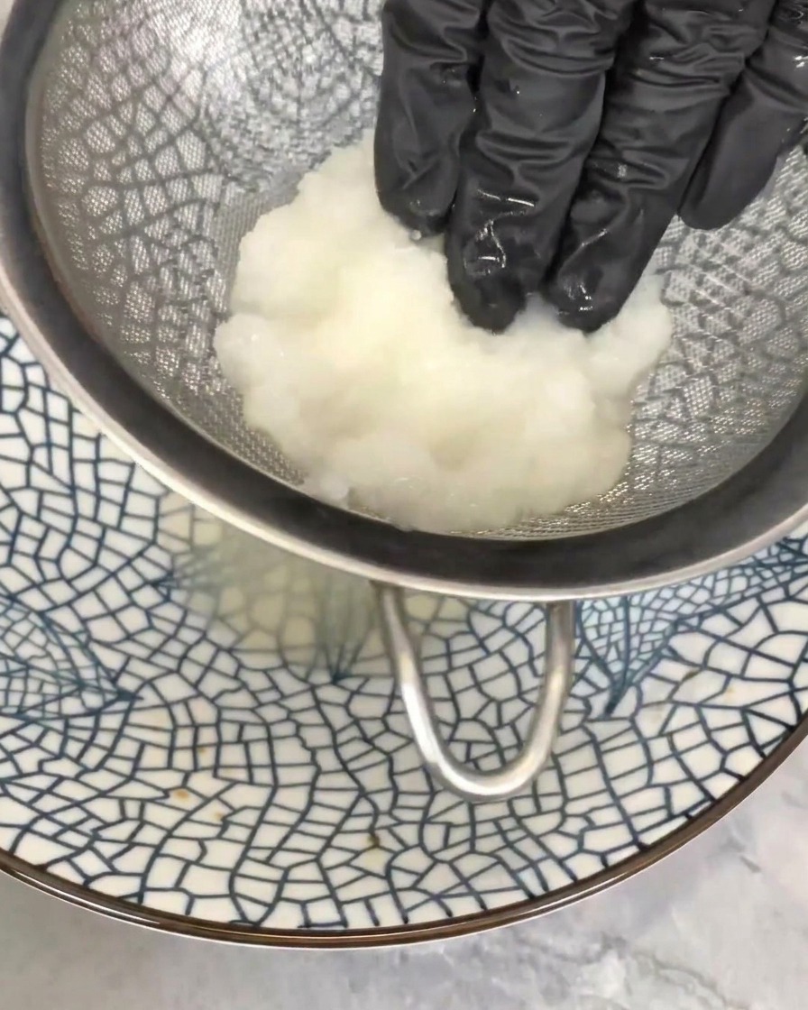 A gloved hand pressing a pile of white grated radish into a mesh sieve over a bowl to drain away the excess liquid.