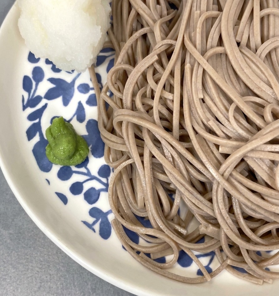 A close-up of chilled buckwheat noodles served on a blue and white floral plate, garnished with a small portion of green wasabi and a white, molded ball of grated radish.