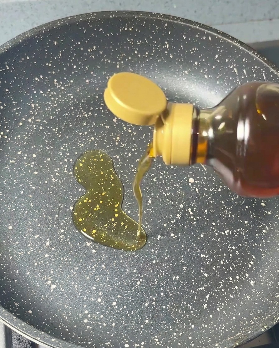 Sesame oil being poured from a bottle into a dark, speckled frying pan in a steady circular motion.
