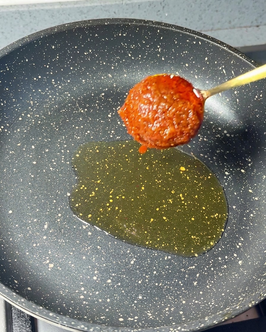 A spoonful of thick red shrimp paste being lowered into a frying pan with a small pool of sesame oil.