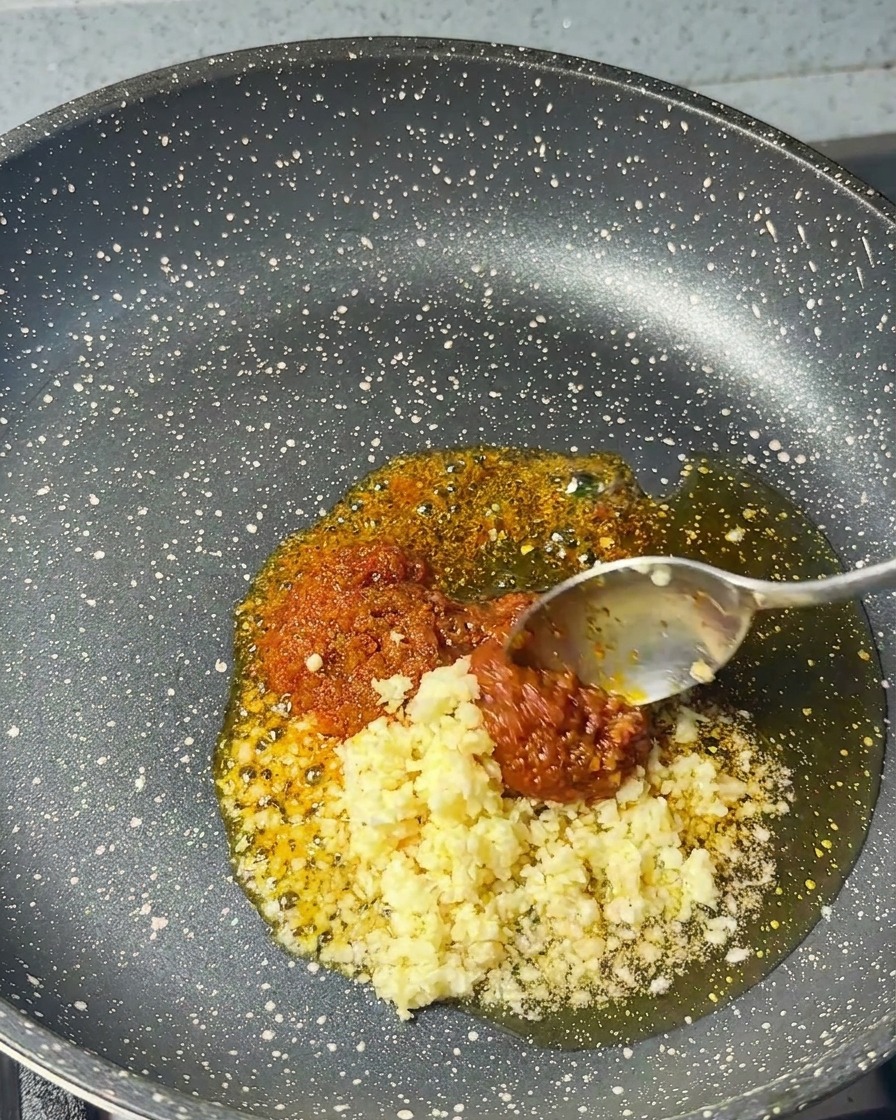 Minced garlic being added from a spoon into a pan already containing red shrimp paste and sesame oil.