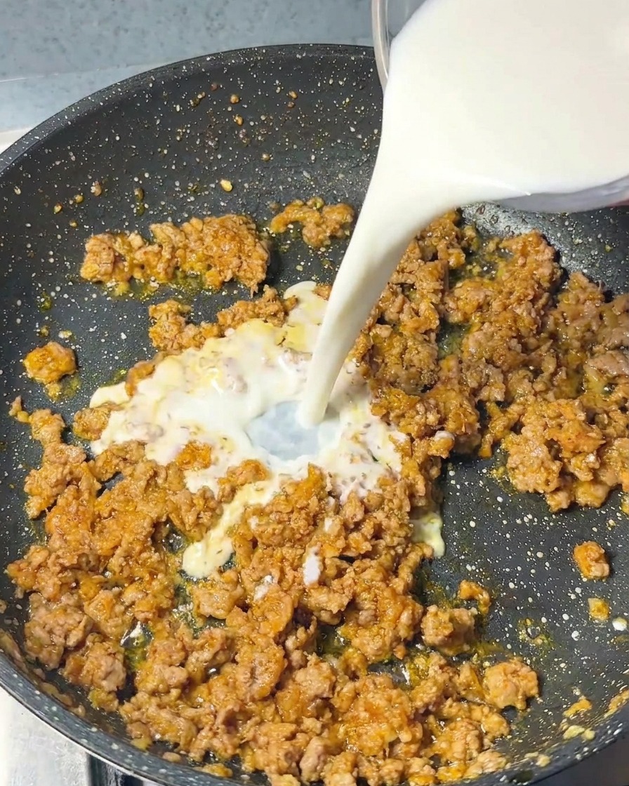 A stream of white milk being poured from a glass container into a frying pan filled with cooked ground pork and seasonings.