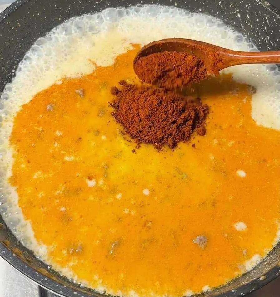 A wooden spoon adding pieces of curry roux into a bubbling milk-based sauce in a dark pan.