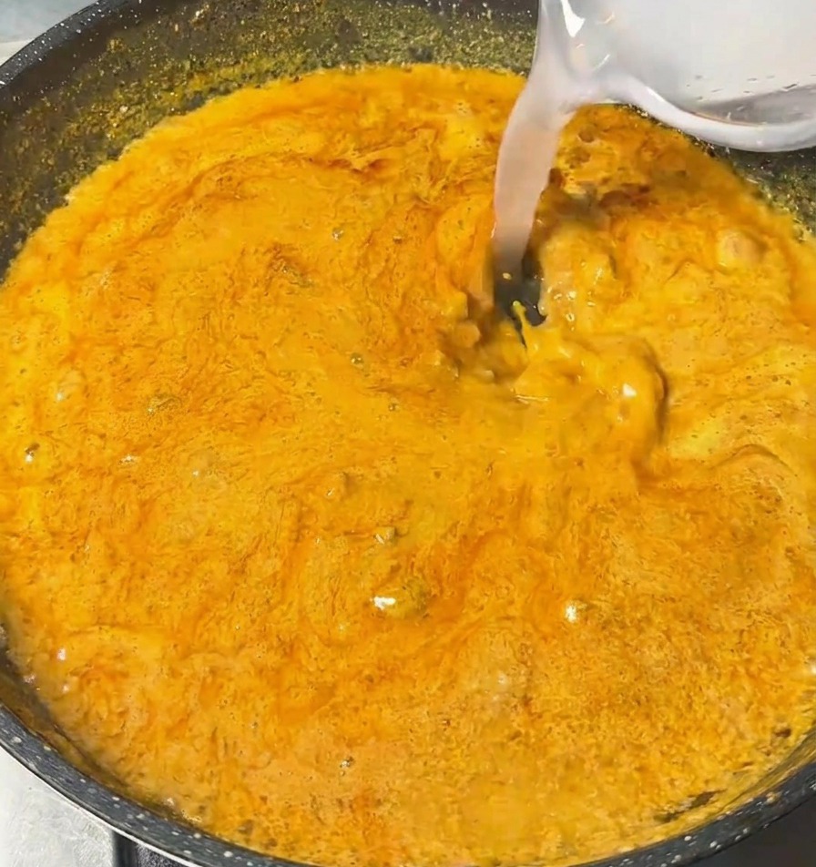 A clear liquid being poured from a small bowl into a pan of simmering, thick orange curry sauce.