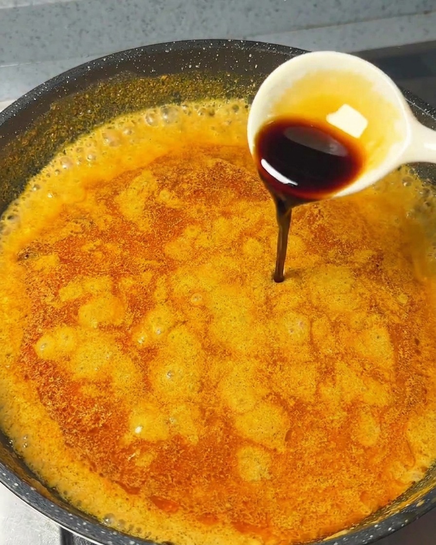 A white spoon pouring dark soy sauce into a bubbling orange curry sauce in a pan.