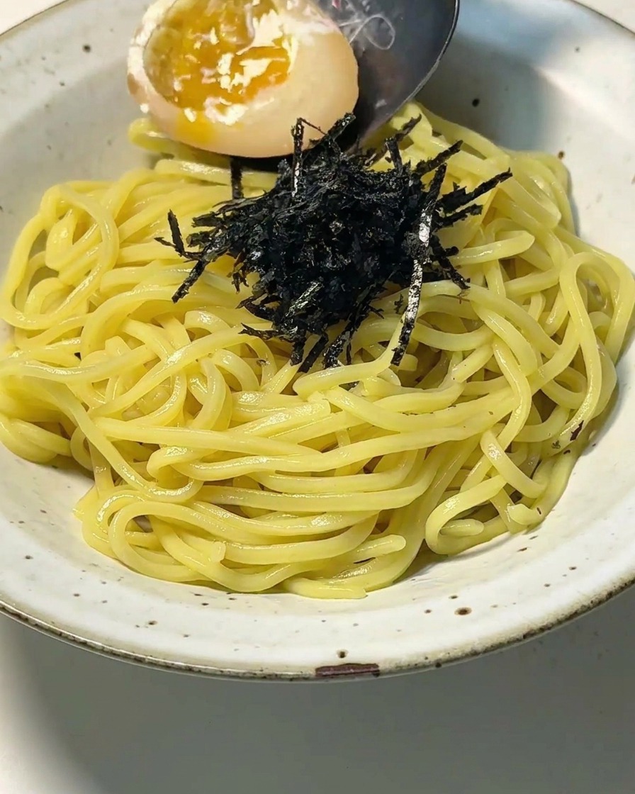 A close-up view of a white ceramic bowl filled with yellow ramen noodles and shredded nori, as a spoon places a halved soft-boiled egg with a glistening, runny yolk onto the dish.