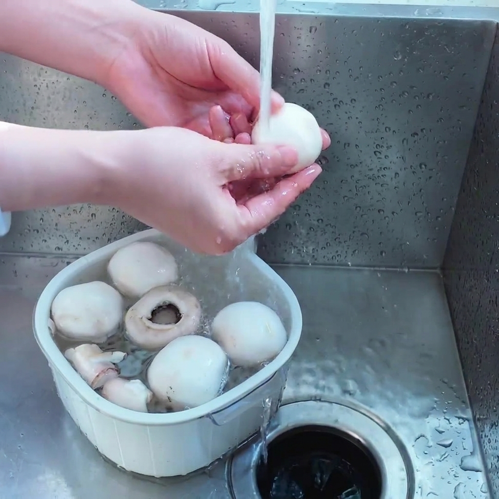 A person washing several whole white button mushrooms in a small white plastic strainer under a steady stream of tap water.