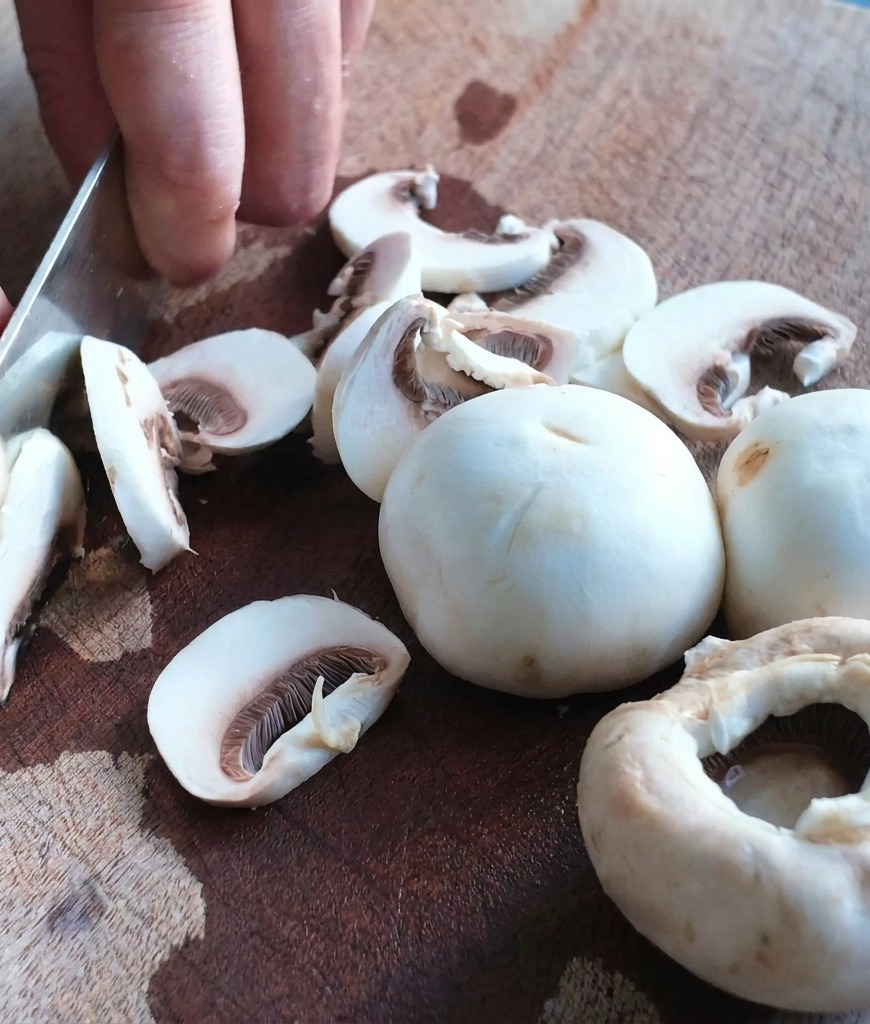 Close-up of a metal chefs knife slicing through white mushrooms on a textured wooden cutting board.